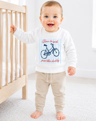 A smiling baby boy wearing a white sweatshirt with a bicycle graphic and the text "Born to cycle just like daddy" is standing next to a wooden crib.