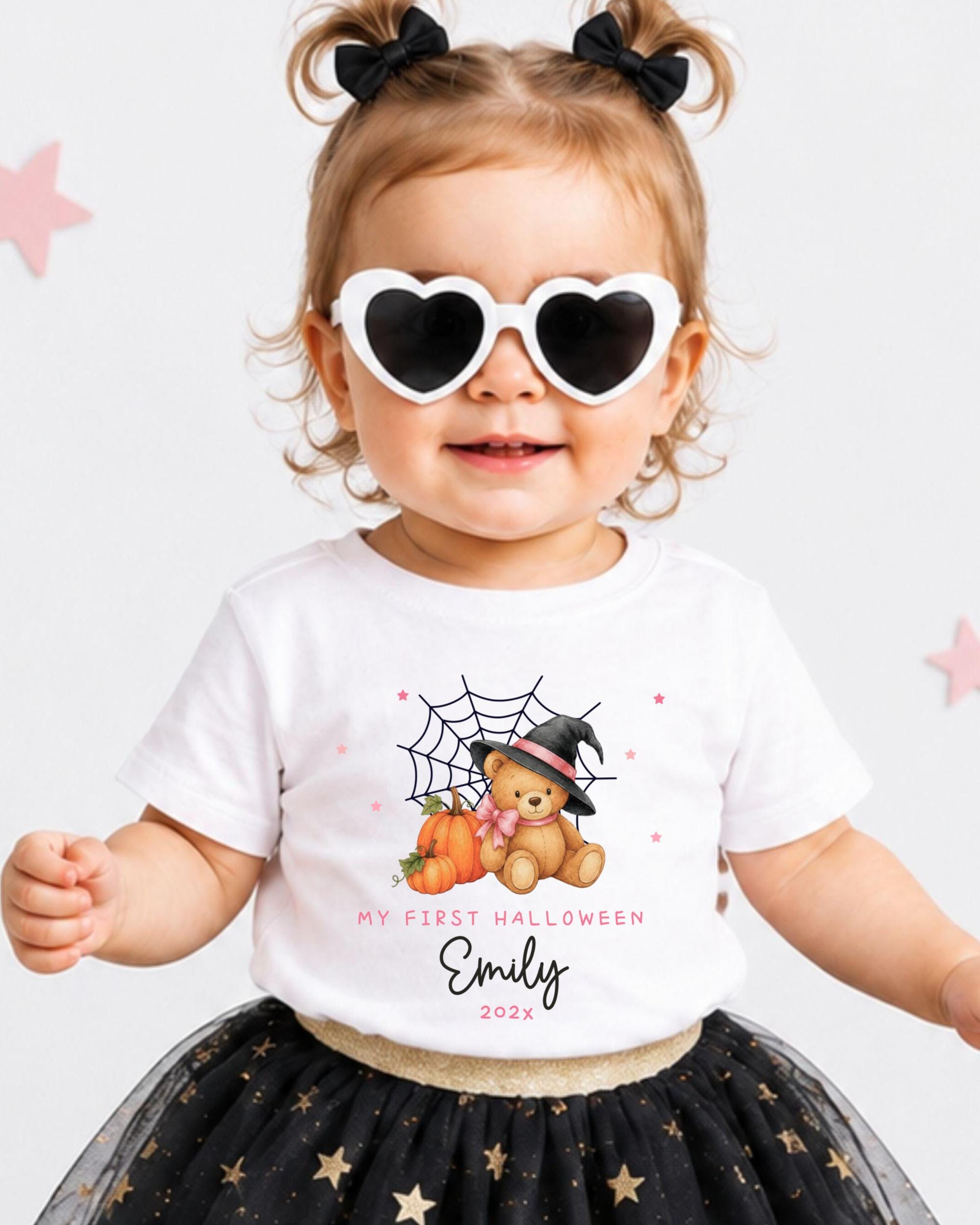 A young girl wearing a white t-shirt with a Halloween-themed design, including a teddy bear, spider web, and pumpkins, along with heart-shaped sunglasses and a black and gold tutu skirt.