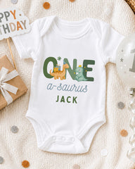 A white onesie with the text "ONE a-Saurus JACK" printed on it, along with illustrations of a dinosaur and a dinosaur. The onesie is laid out on a blanket, surrounded by a birthday card, a balloon, and confetti.