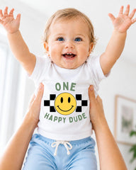A smiling baby wearing a white shirt with a smiley face and the text "ONE HAPPY Dude" is being held up by someone's arms.
