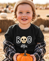 A young child wearing a black t-shirt with a skull design and the text "BOO" and "Alfie" is holding a small orange pumpkin.