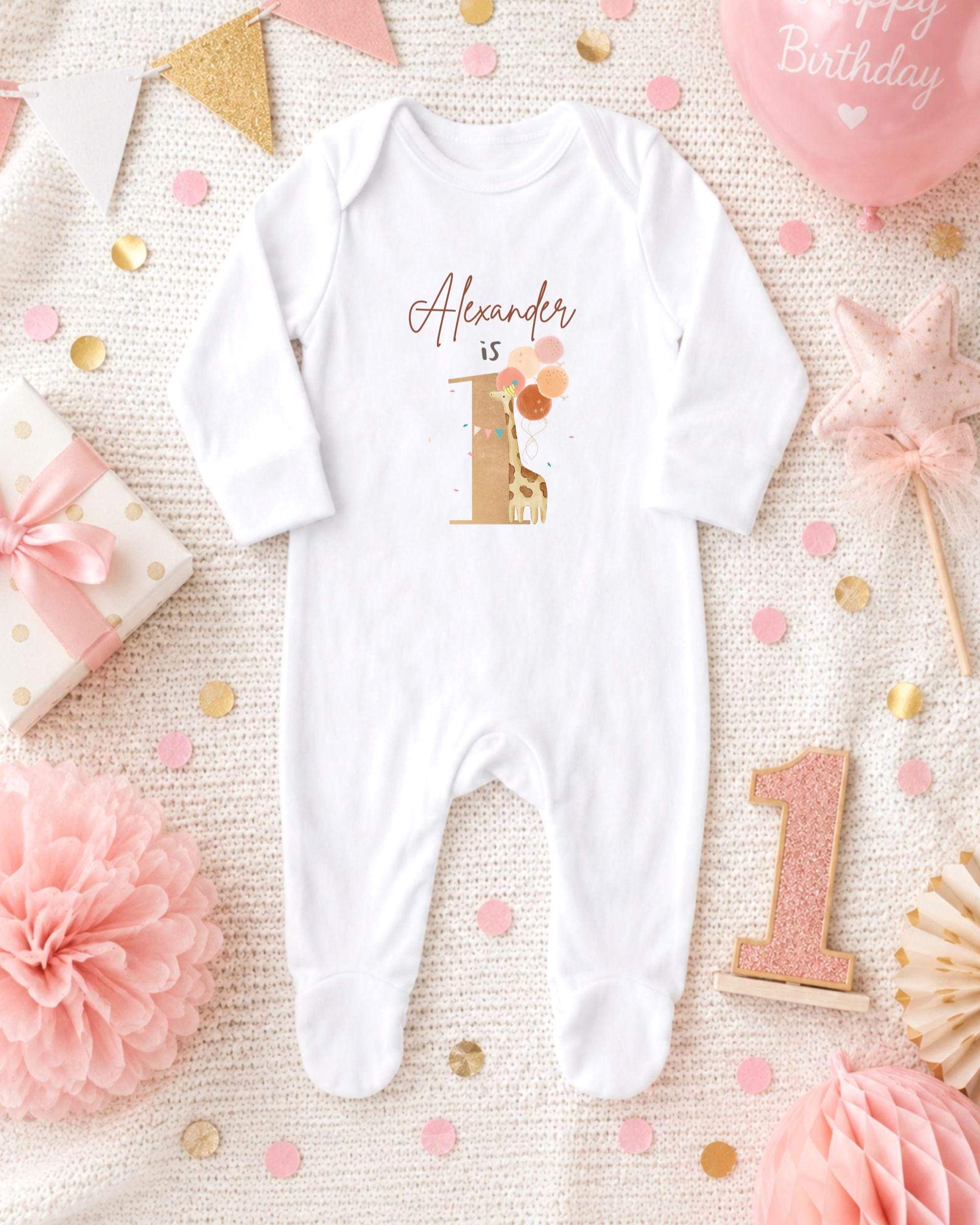 A white onesie with the name "Alexander" printed on it, surrounded by pink and gold decorations, confetti, and a pink balloon.
