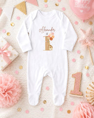 A white onesie with the name "Alexander" printed on it, surrounded by pink and gold decorations, confetti, and a pink balloon.