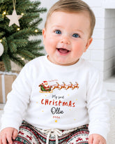 A smiling baby wearing a white shirt with a Christmas-themed design and text, sitting in front of a Christmas tree.