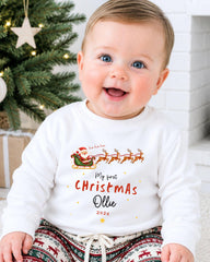 A smiling baby wearing a white shirt with a Christmas-themed design and text, sitting in front of a Christmas tree.