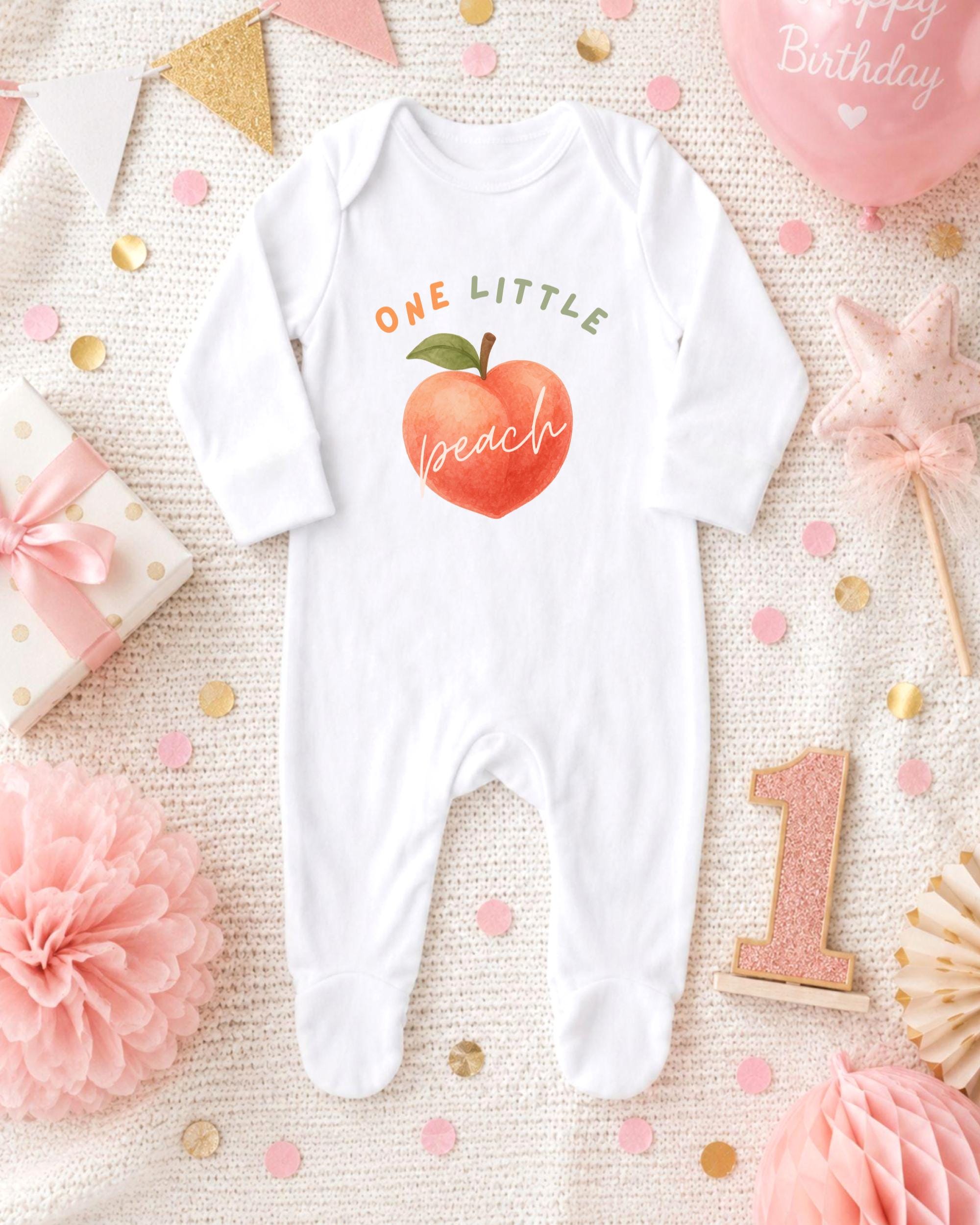 A white onesie with a peach design and the text "ONE LITTLE PEACH" is displayed on a bed with confetti, a pink star, and other baby-related items.