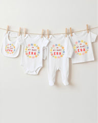 A row of four white baby clothes with the text "IN MY BABY ERA" printed on them, hanging on a clothesline.
