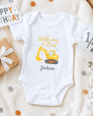 A white onesie with a yellow construction vehicle graphic and the text "Halfway to One Jackson" printed on it, along with a "Happy Birthday" sign and a gift box.