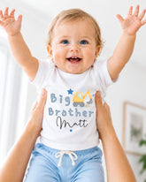 A smiling baby wearing a white shirt with a construction vehicle design and the text "Big Brother Matt" is being held up by someone's arms.