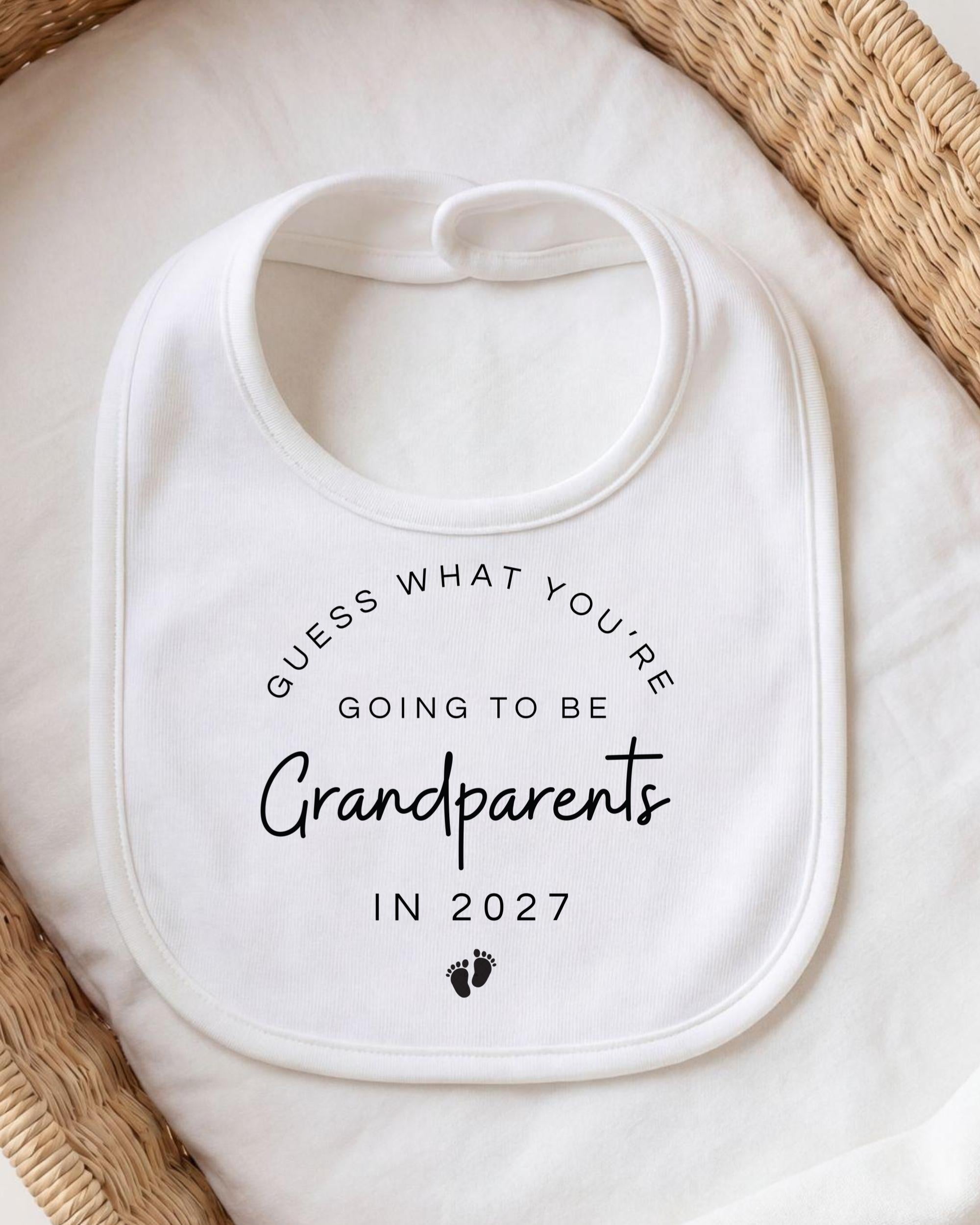 A white bib with text that says "Guess what you're going to be Grandparents in 2027" printed on it.