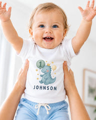 A smiling baby wearing a white t-shirt with a dinosaur graphic and the name "Johnson" printed on it, being held up by someone's arms.