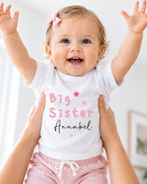 A smiling baby girl wearing a white shirt with the text "Big Sister Annabel" is being held up by someone's arms.