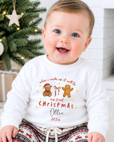 A smiling baby wearing a white long-sleeved shirt with a Christmas-themed design and text.