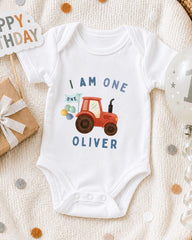 A white onesie with a red tractor design and the text "I AM ONE OLIVER" printed on it, along with a birthday sign and a balloon.
