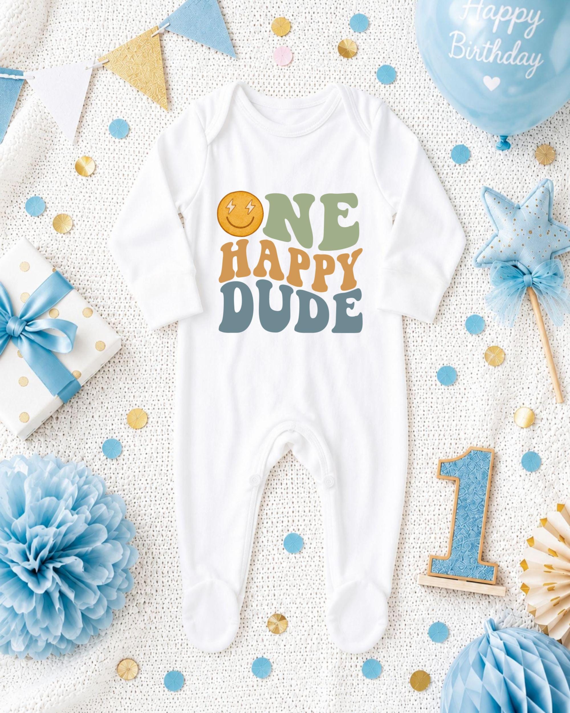 A white onesie with the text "ONE HAPPY DUDE" printed on it, surrounded by birthday decorations such as a blue balloon, confetti, and a star-shaped ornament.