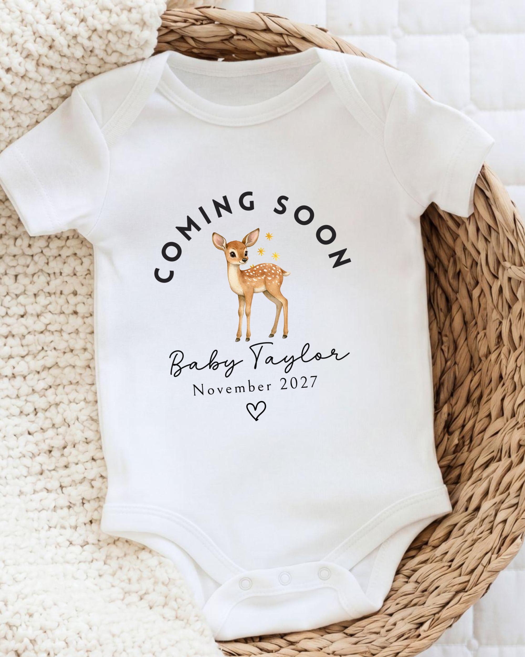 A white baby onesie with a graphic of a deer and the text "Coming soon Baby Taylor November 2027" printed on it.