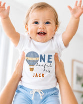 A smiling baby wearing a white shirt with a hot air balloon and the text "One derful boy Jack" is being held up by someone's arms.