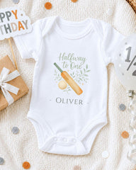 A white onesie with a cricket bat and ball design, along with a "Happy Birthday" sign and confetti, is displayed on a white blanket.