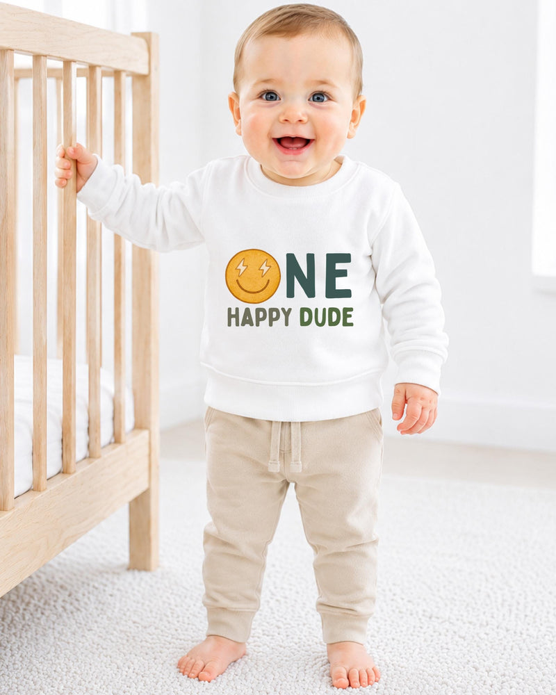 A smiling baby boy wearing a white sweatshirt with a smiley face and the text "ONE HAPPY Dude" is standing next to a wooden crib.