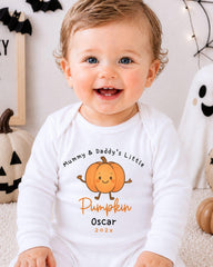 A smiling baby boy wearing a white onesie with a pumpkin graphic and text, sitting in front of Halloween decorations.