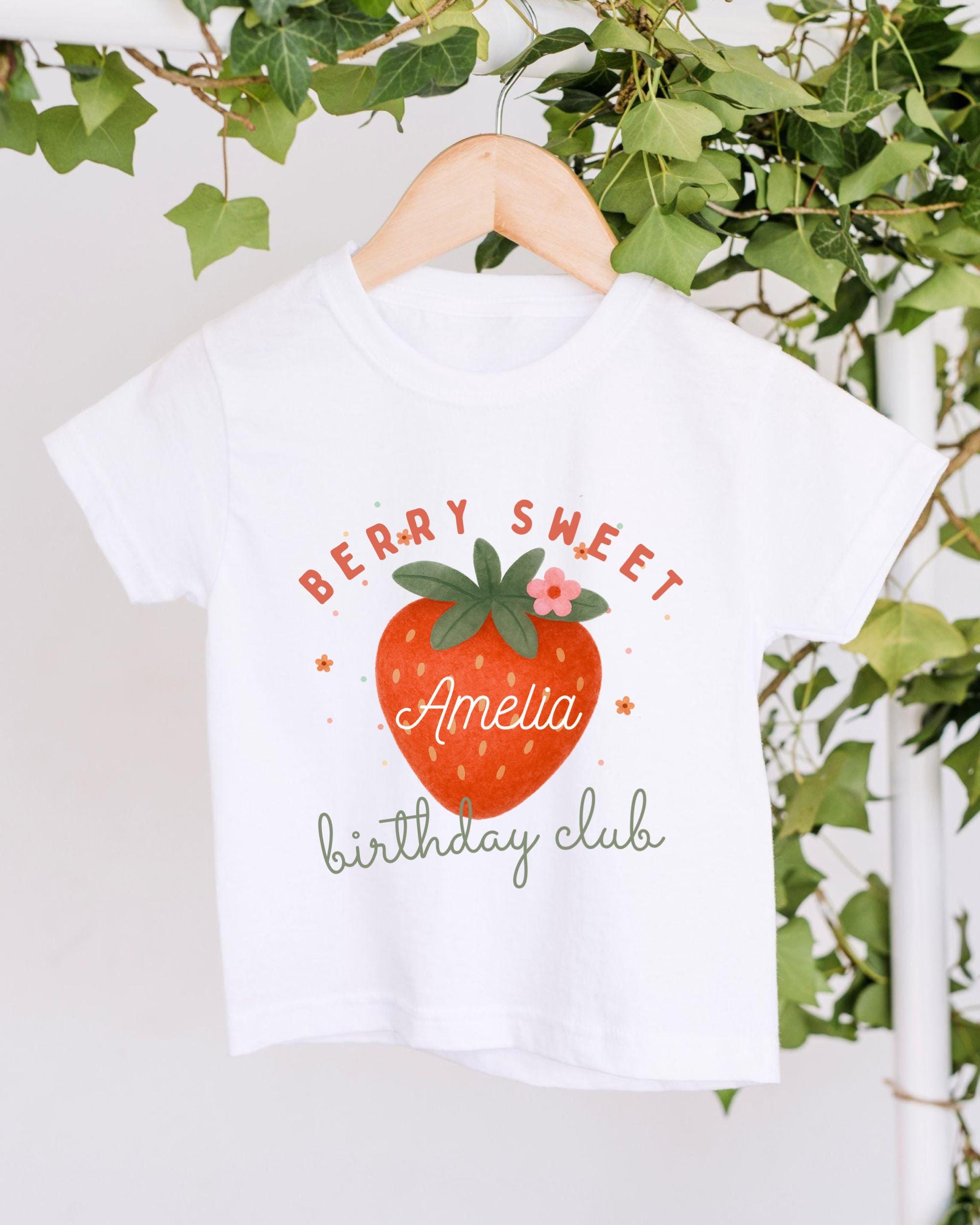 A white t-shirt with a graphic design featuring a red strawberry and the text "Berry Sweet Amelia birthday club" hanging on a wooden hanger.