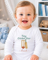A smiling baby boy wearing a white onesie with a cricket-themed design, sitting in a room with a teddy bear and bookshelf in the background.