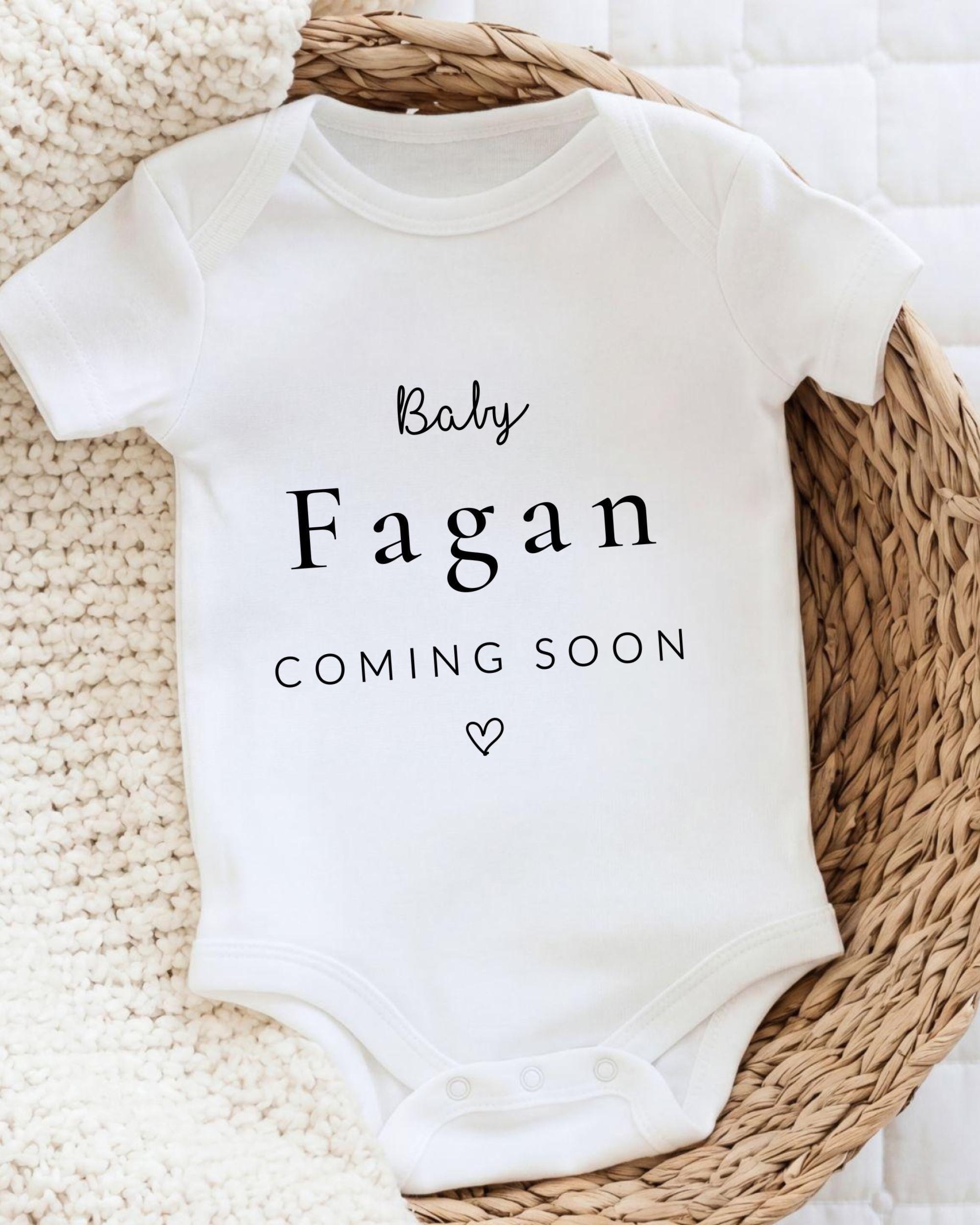 A white baby onesie with the text "Baby Fagan Coming soon" printed on it, placed on a woven basket.
