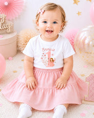 A smiling baby girl wearing a white t-shirt with the number 1 and a pink skirt, sitting on a pink and white background with balloons and confetti.