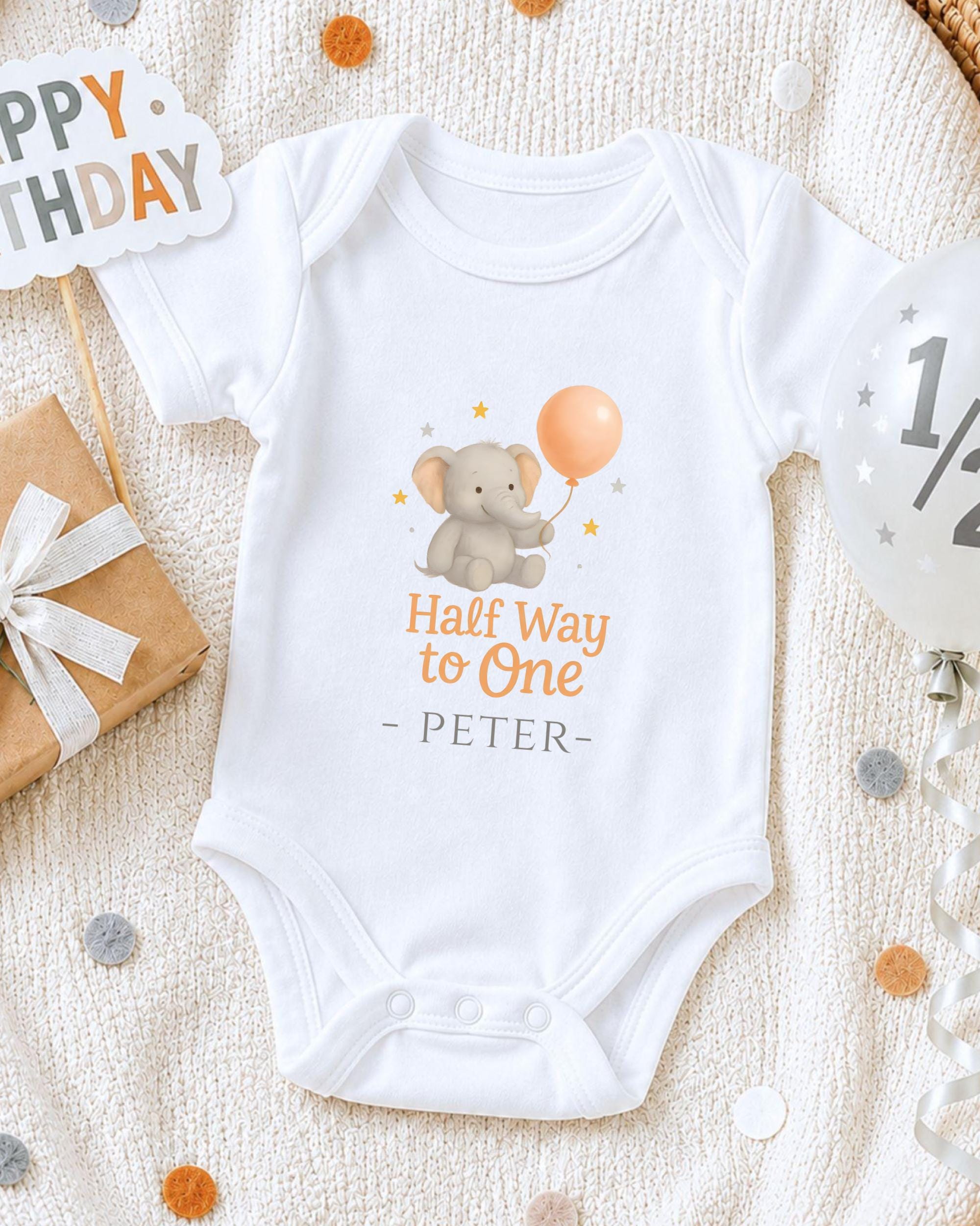 A white onesie with a cartoon elephant and the text "Half Way to One - Peter" printed on it, along with a "Happy Birthday" sign and confetti.