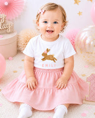 A smiling baby girl wearing a white t-shirt with a bunny graphic and pink skirt, sitting on a pink and white background with balloons and confetti.