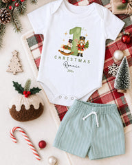 A white onesie with a Christmas-themed design, a pair of green shorts, and various Christmas decorations such as a small Christmas tree, ornaments, and a nutcracker.