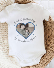 A white baby onesie with a heart-shaped photo of a man and a child, along with the text "Loved & Protected by my Grandpa in Heaven" printed on it.
