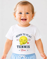 A smiling baby boy wearing a white t-shirt with a tennis-themed design.