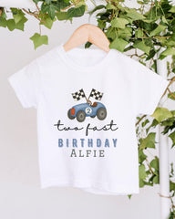 A white t-shirt with a graphic of a race car and the text "two fast birthday alfie" printed on it, hanging on a wooden hanger.