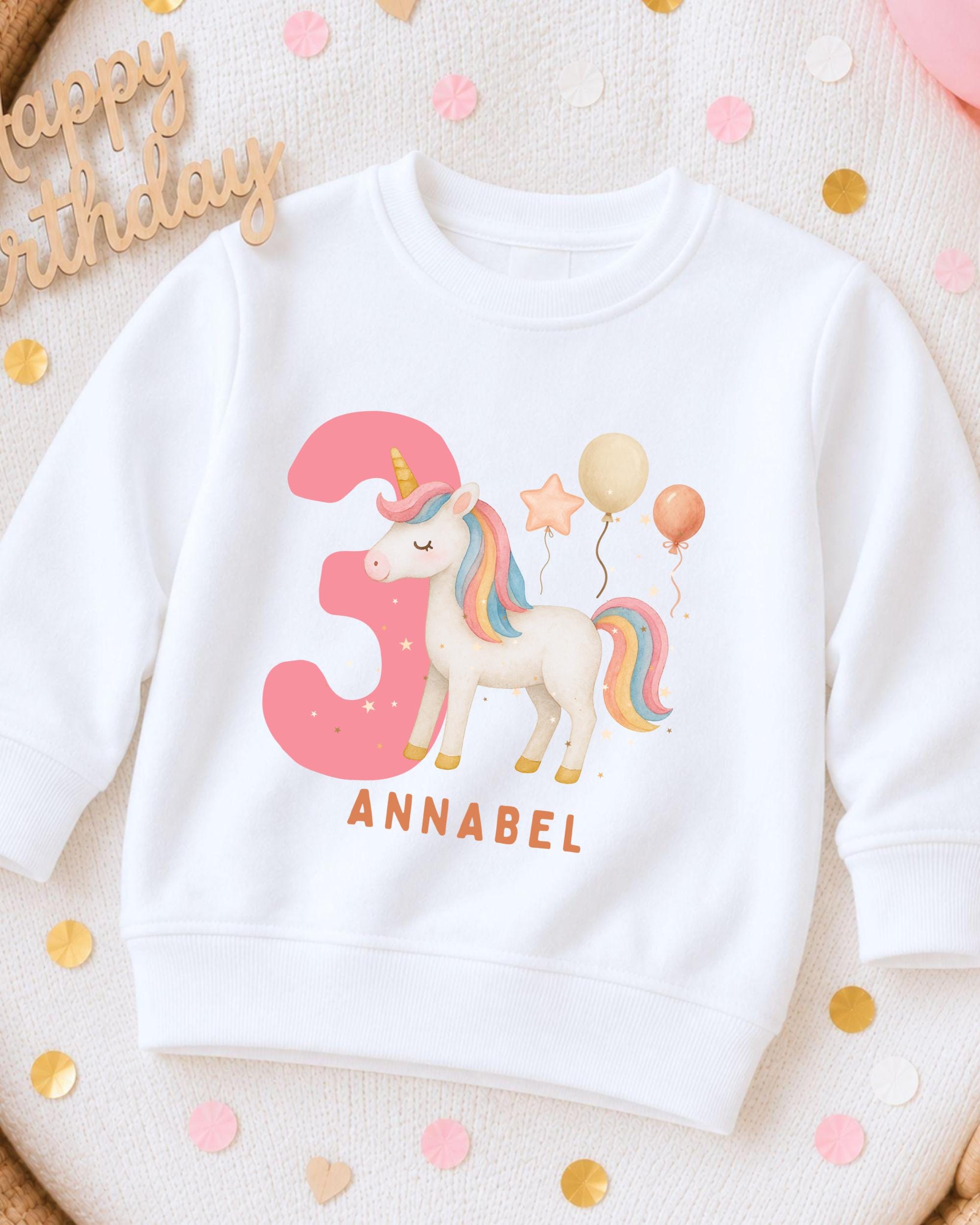 A white sweatshirt with a unicorn and balloons printed on it, along with the name 'Annabel' printed below.