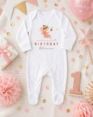 A white onesie with a fairy design and the text "my fairy first birthday Olivia" printed on it, surrounded by pink and gold decorations, a pink star, and a pink and gold birthday cake.