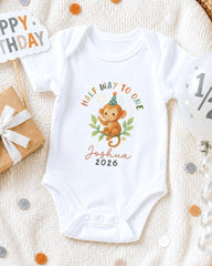 A white onesie with a monkey graphic and the text "Half Way to One" printed on it, along with the name "Joshua" and the year "2026". The onesie is placed on a white blanket, surrounded by birthday decorations such as a "Happy Birthday" sign, a balloon, and confetti.