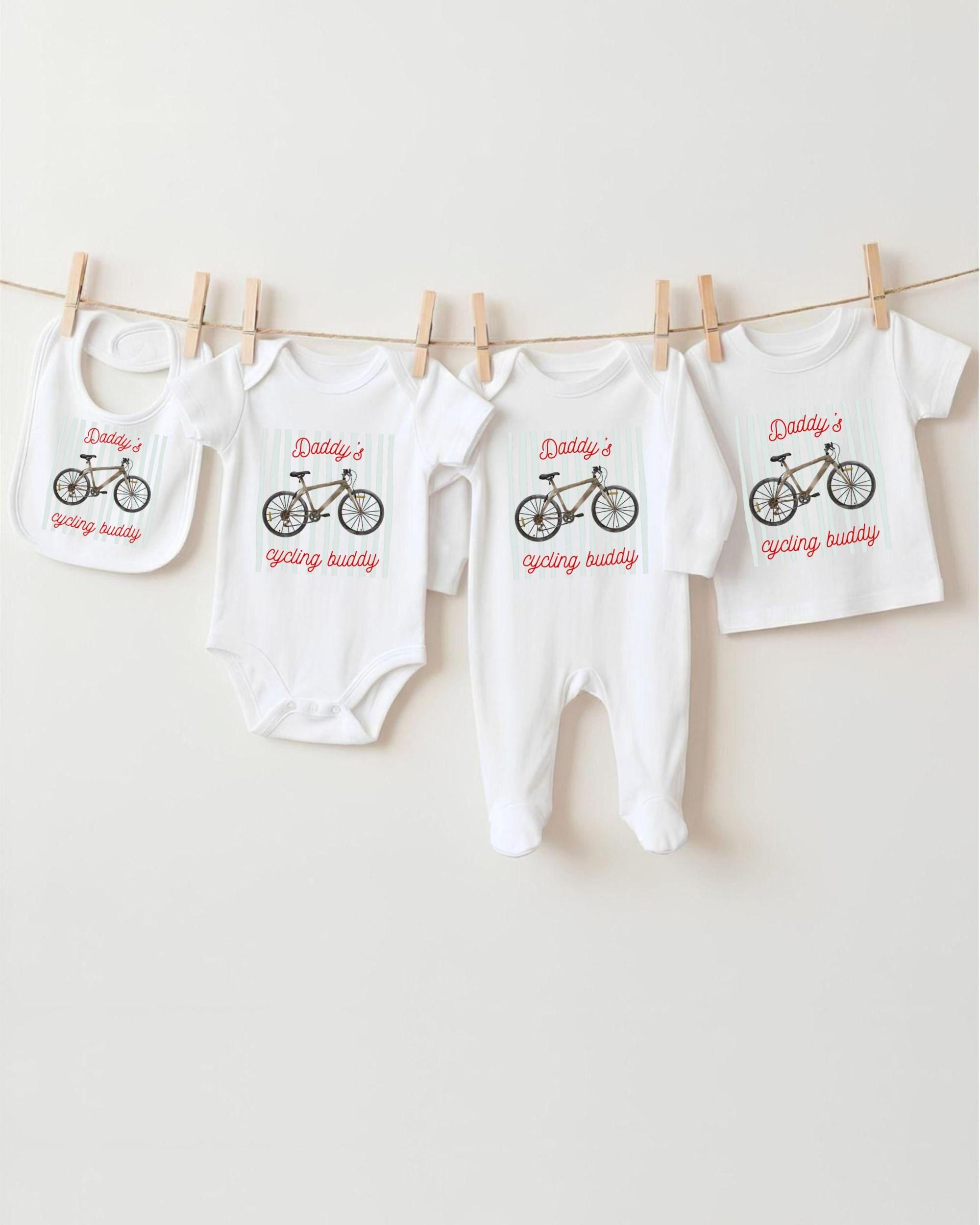 A row of four white baby clothes with bicycle designs, each featuring the text "Daddy's cycling buddy" printed on them.
