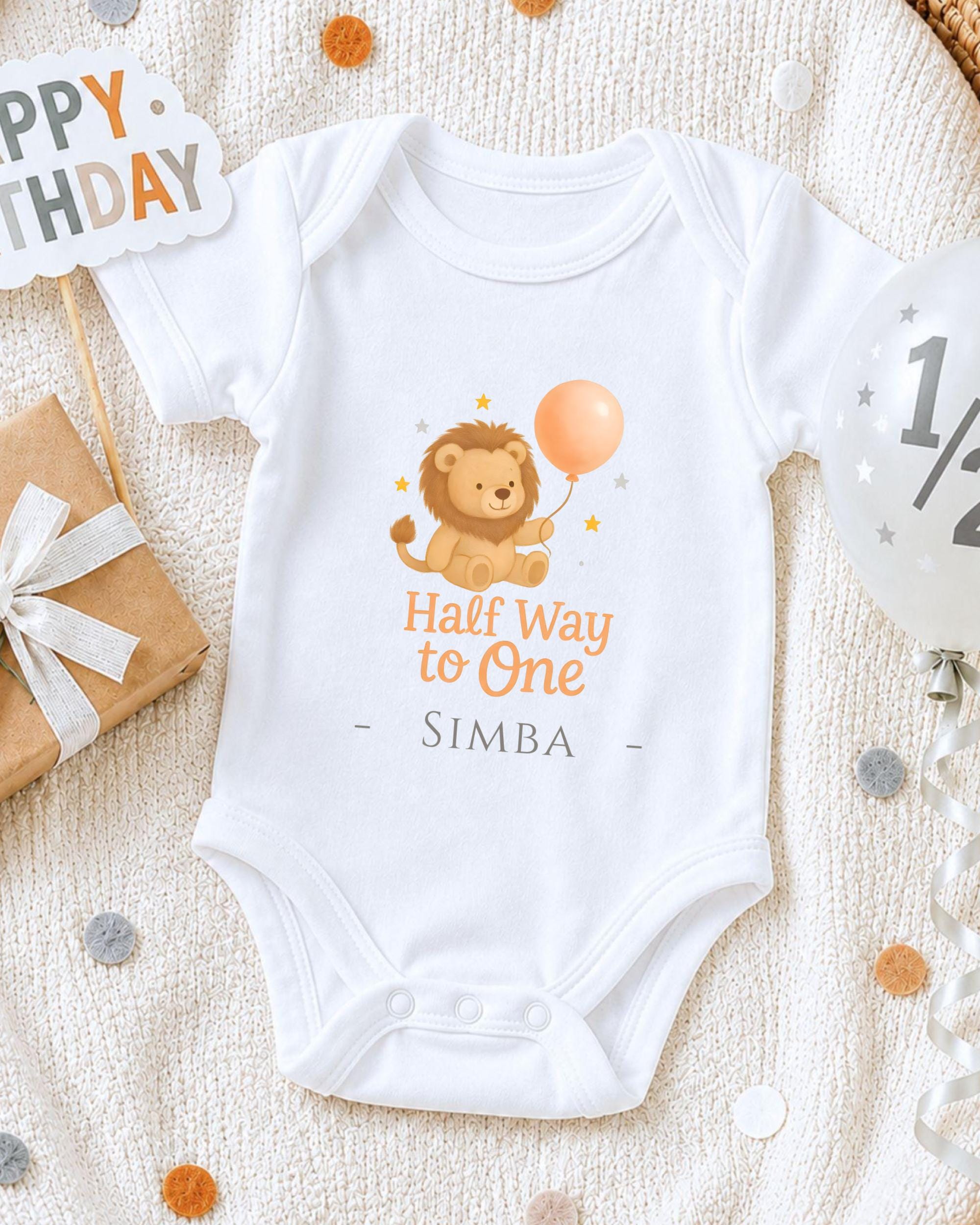A white onesie with a cartoon lion and a balloon, along with a "Happy Birthday Simba" sign, is displayed on a white blanket.