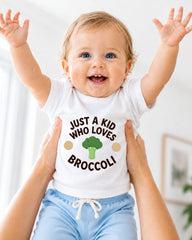 A smiling baby wearing a white shirt with the text "Just a kid who loves broccoli" is being held up by someone's arms.