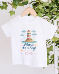 A white t-shirt with a lighthouse graphic and the text "Ahoy, it's a boy!" printed on it, hanging on a wooden hanger.