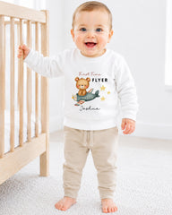 A smiling baby boy wearing a white sweatshirt with a teddy bear and airplane design, standing in a nursery setting.