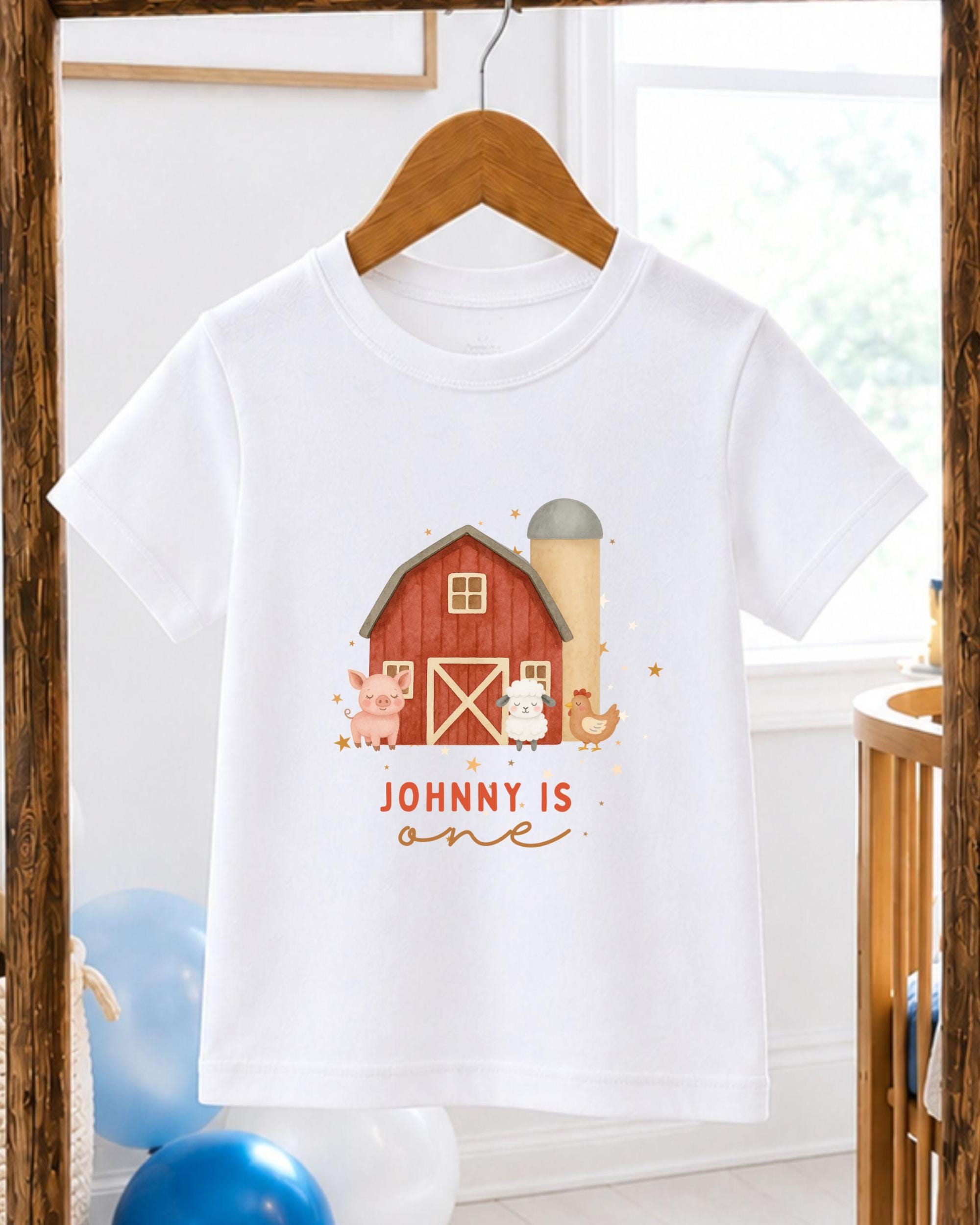 A white t-shirt with a graphic design of a red barn and farm animals, including a pig, chicken, and cow, along with the text "JOHNNY IS ONE".