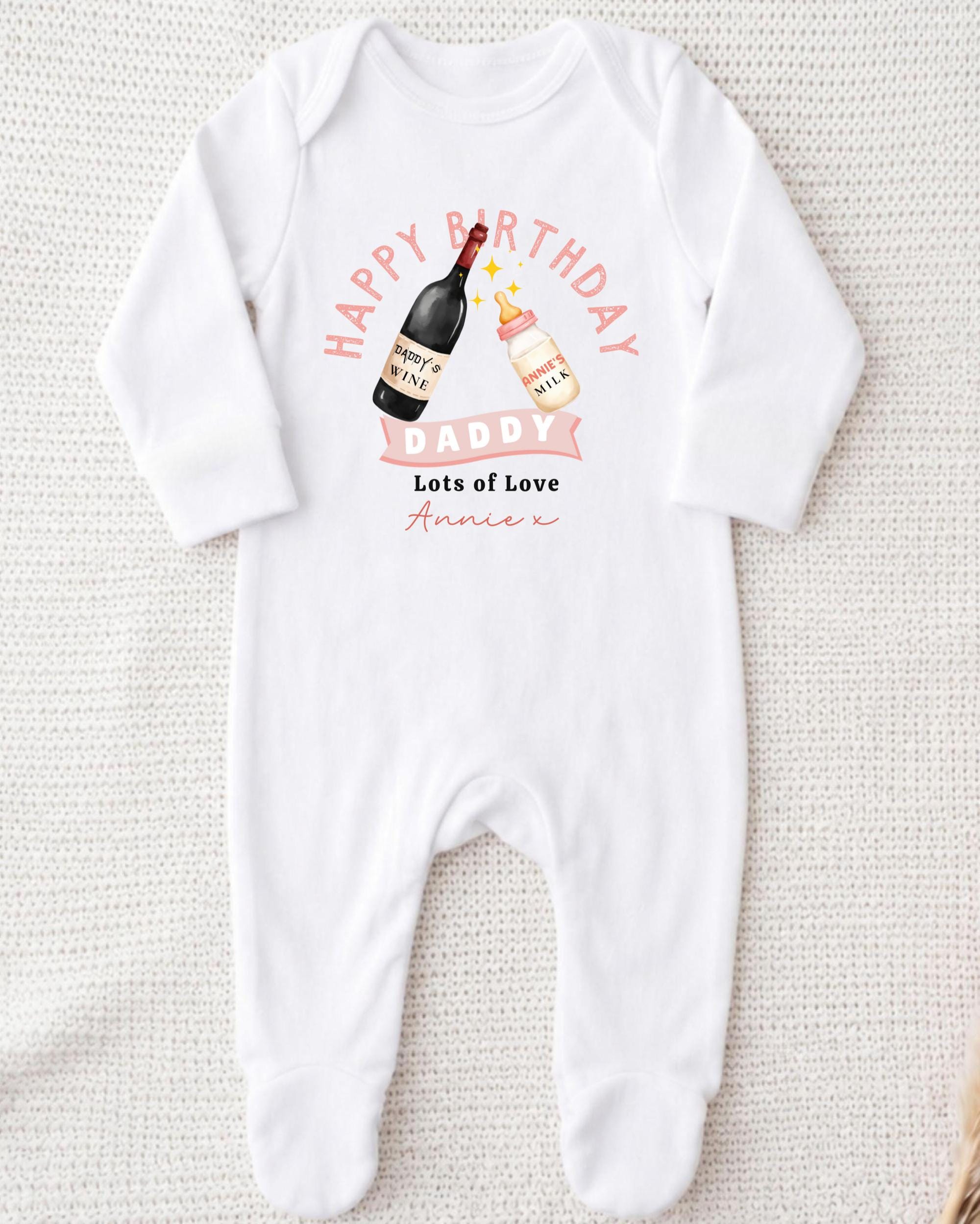 A white onesie with a graphic design on it, featuring a bottle of wine and a baby bottle, along with the text "Happy Birthday Daddy Lots of Love Annie" printed on it.