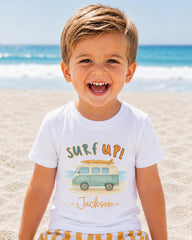A young boy with a big smile on his face, wearing a white t-shirt with a surfboard graphic and the text "Surf up! Jackson" printed on it, standing on a sandy beach with the ocean in the background.
