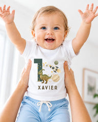 A smiling baby wearing a white t-shirt with a dinosaur graphic and the number 1, being held up by someone's arms.
