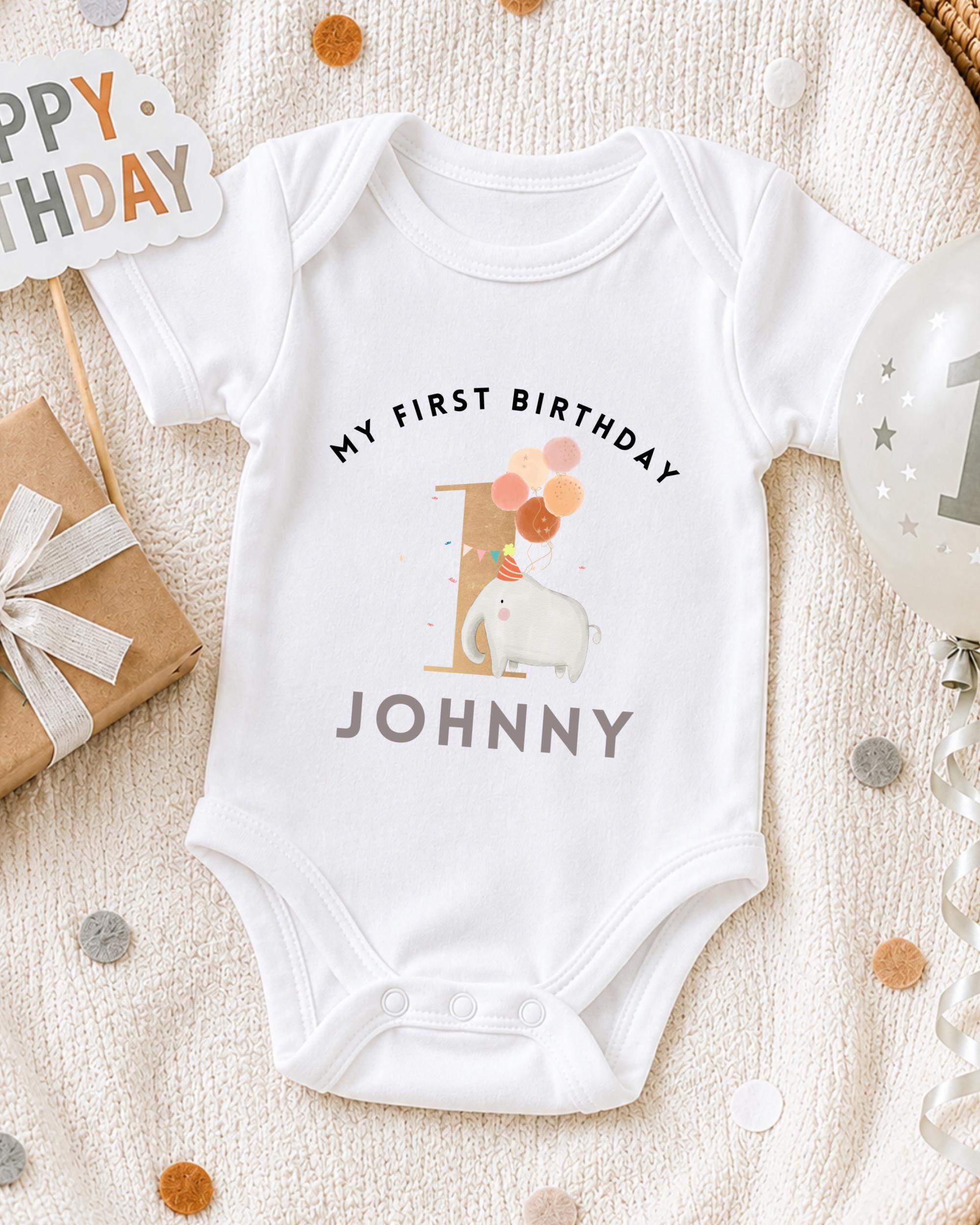 A white onesie with the text "My first birthday Johnny" printed on it, along with an elephant and balloons, is displayed on a bed with a "Happy Birthday" sign and a birthday balloon.