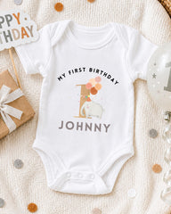 A white onesie with the text "My first birthday Johnny" printed on it, along with an elephant and balloons, is displayed on a bed with a "Happy Birthday" sign and a birthday balloon.