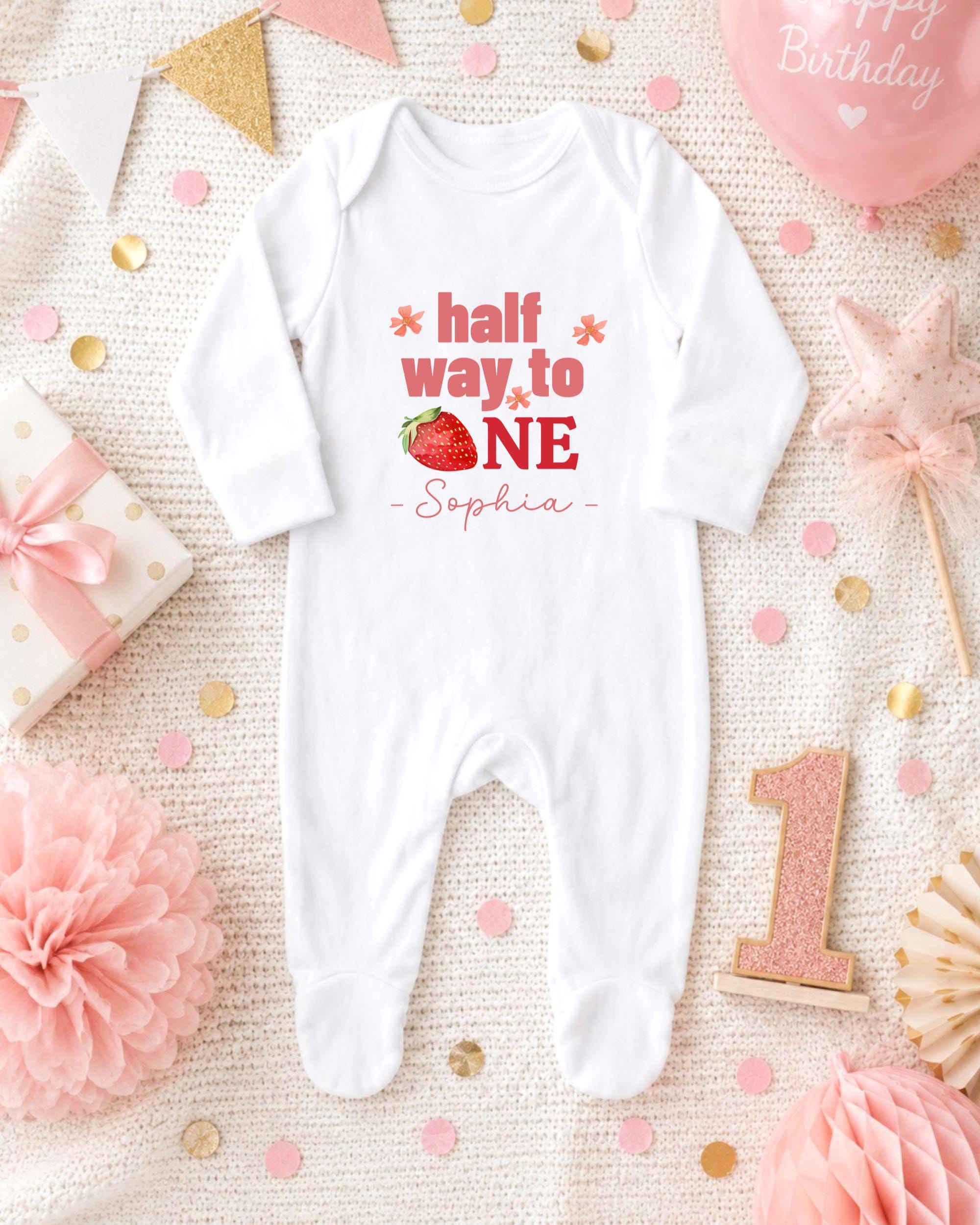 A white onesie with the text "half way to one" printed on it, surrounded by pink and gold confetti, a pink star, and a pink and gold birthday cake.
