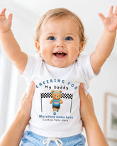 A smiling baby wearing a white t-shirt with a teddy bear and text, being held up by someone's arms.